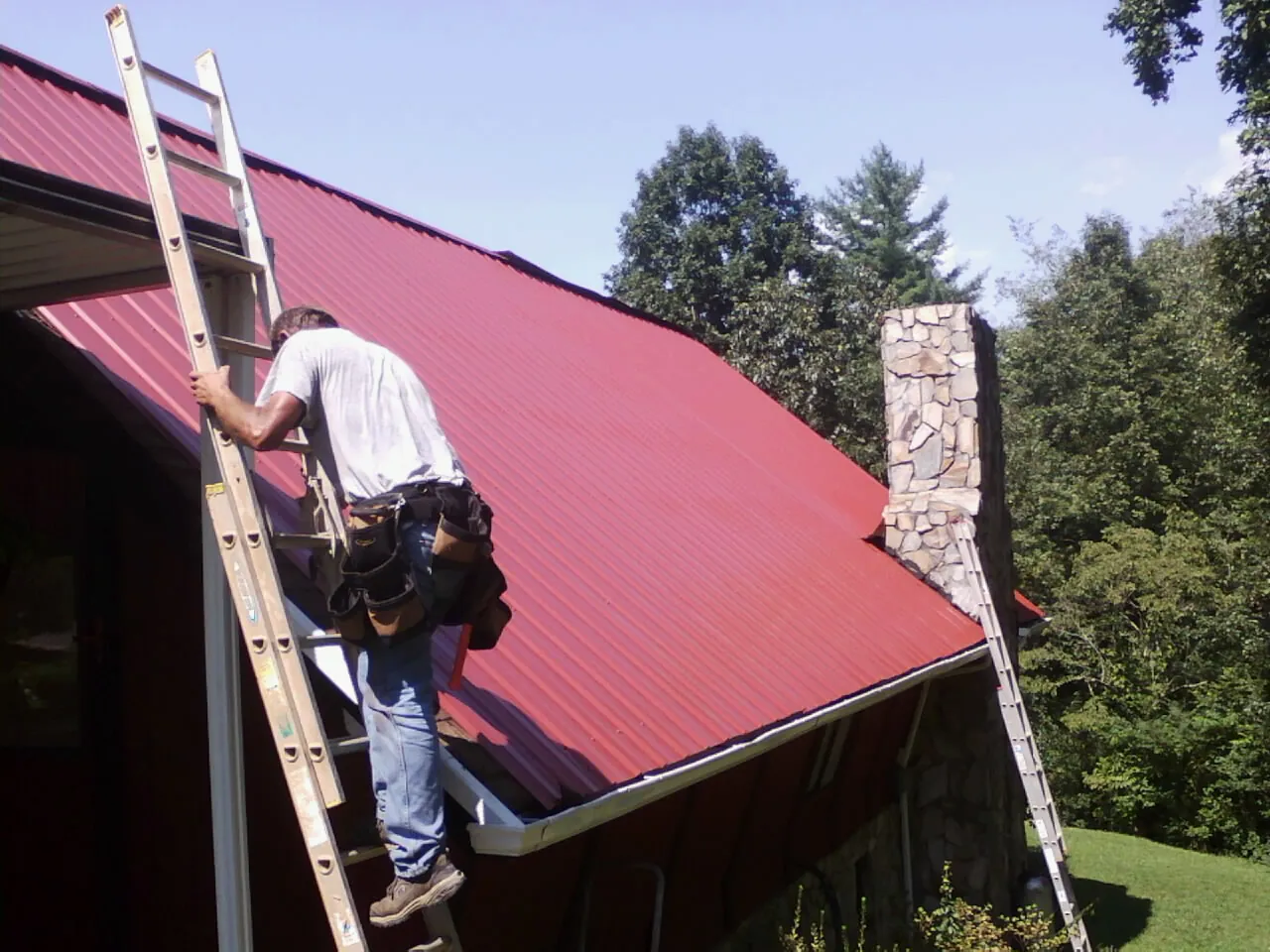 Roofer on a ladder beside a red metal roof and stone chimney — Soffit & Fascia Repair by Roofing Brothers in Hortense